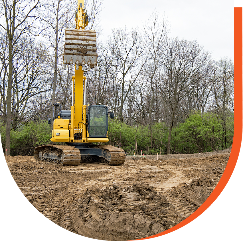 A yellow excavator on muddy ground with bare trees and green shrubs in the background.