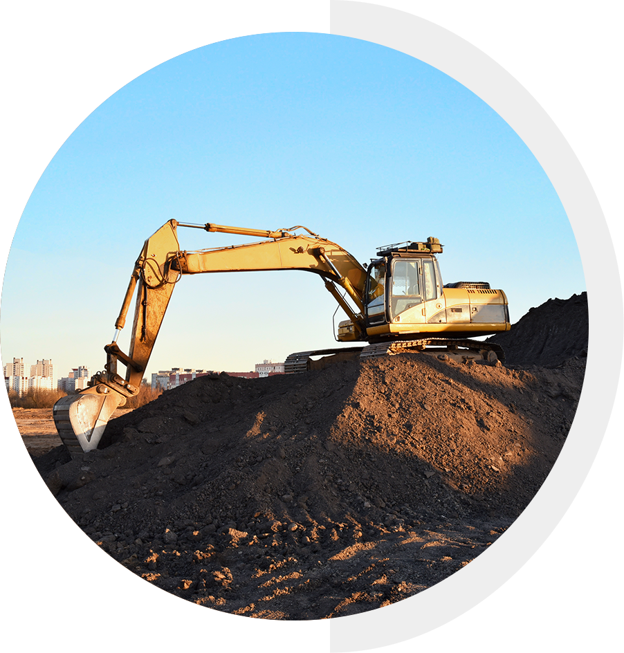 Excavator on a mound of dirt at a construction site, with a city skyline in the background under a clear blue sky.