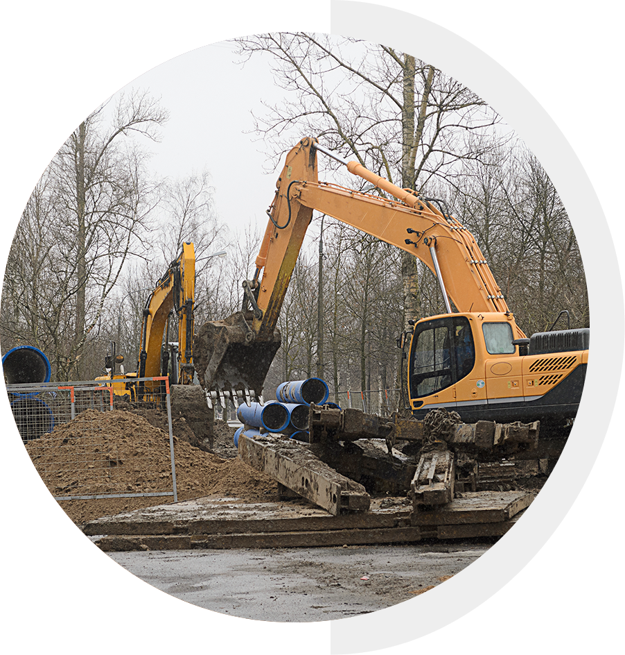 Two excavators are digging on a construction site, surrounded by trees and pipes.