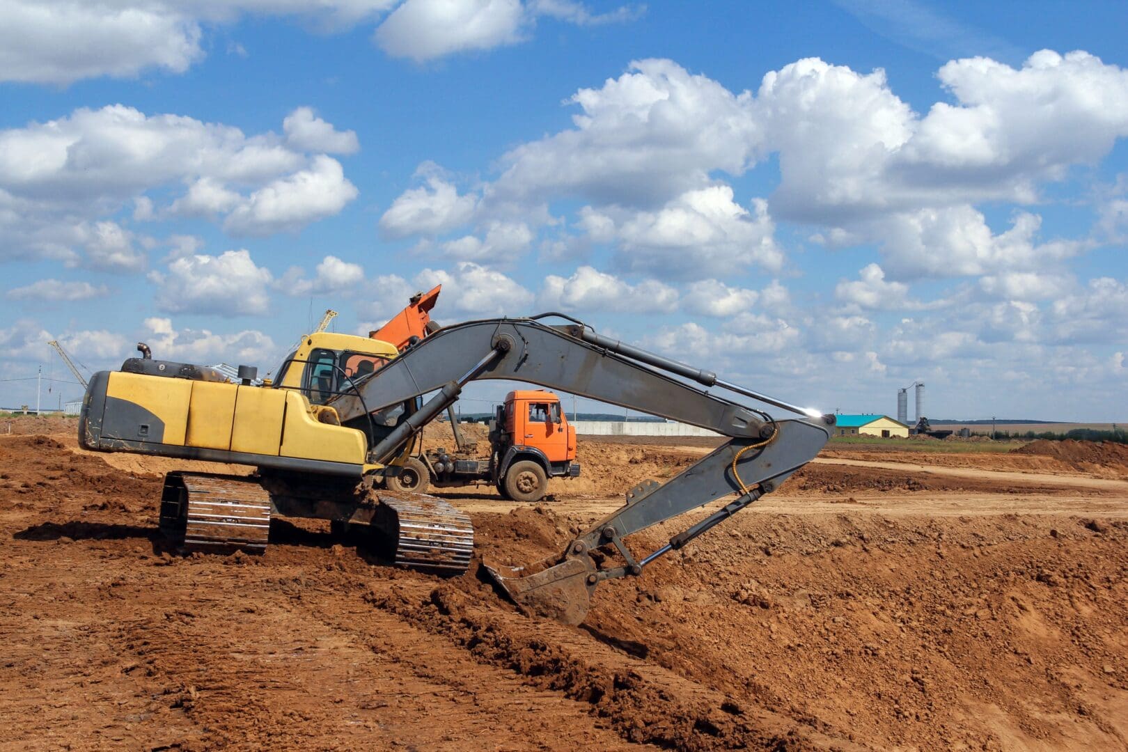 Construction site with a yellow excavator and an orange dump truck working on a dirt landscape under a blue sky with clouds.